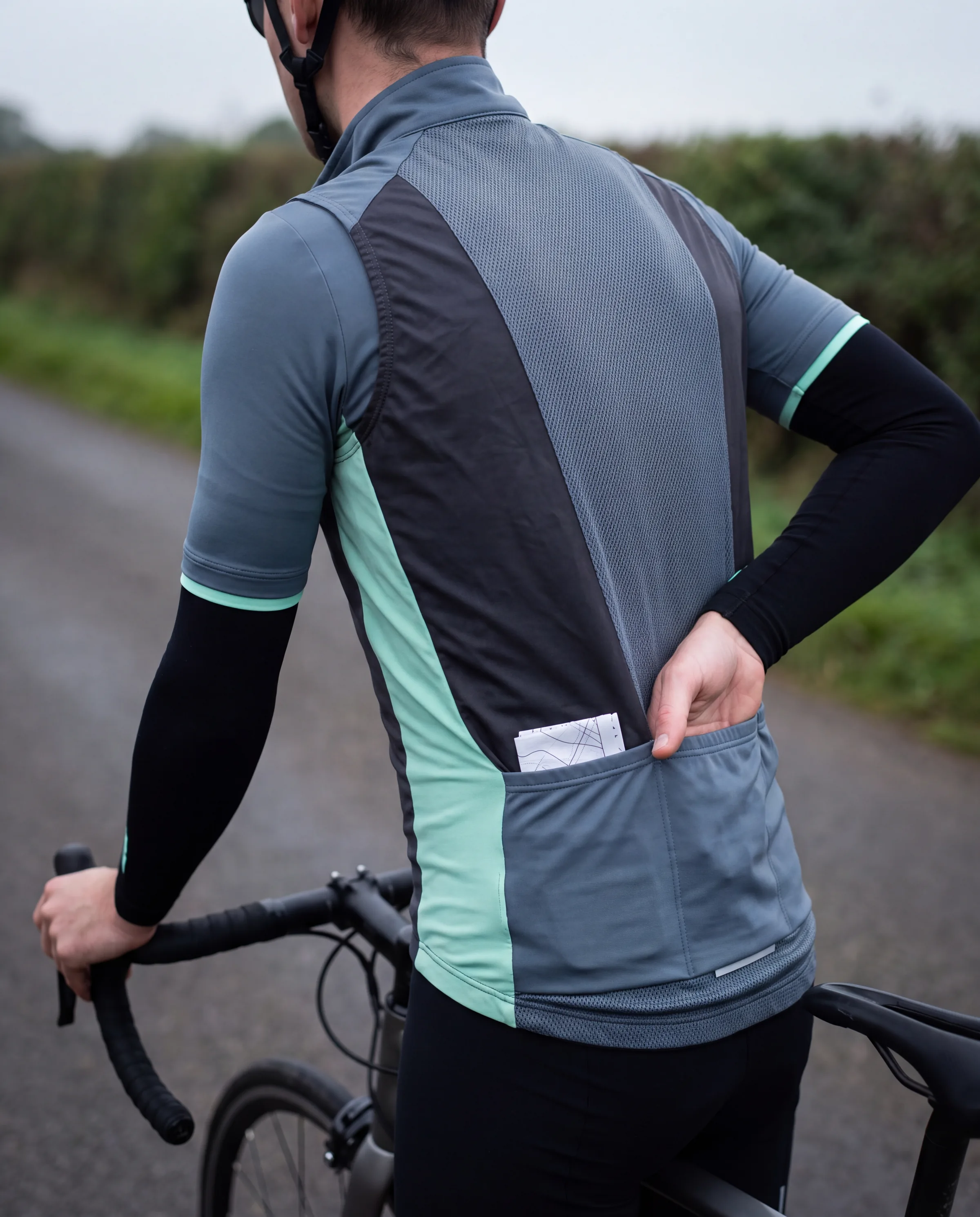 A rider seen from behind reaching under a cycling gilet into the rear pocket of the race jersey beneath, arm warmers under the jersey sleeves